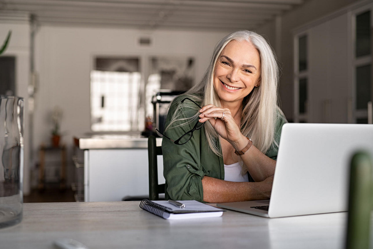 Eine ältere Dame sitzt zu Hause vor ihrem Laptop. Sie hält die Brille in der Hand und lächelt.