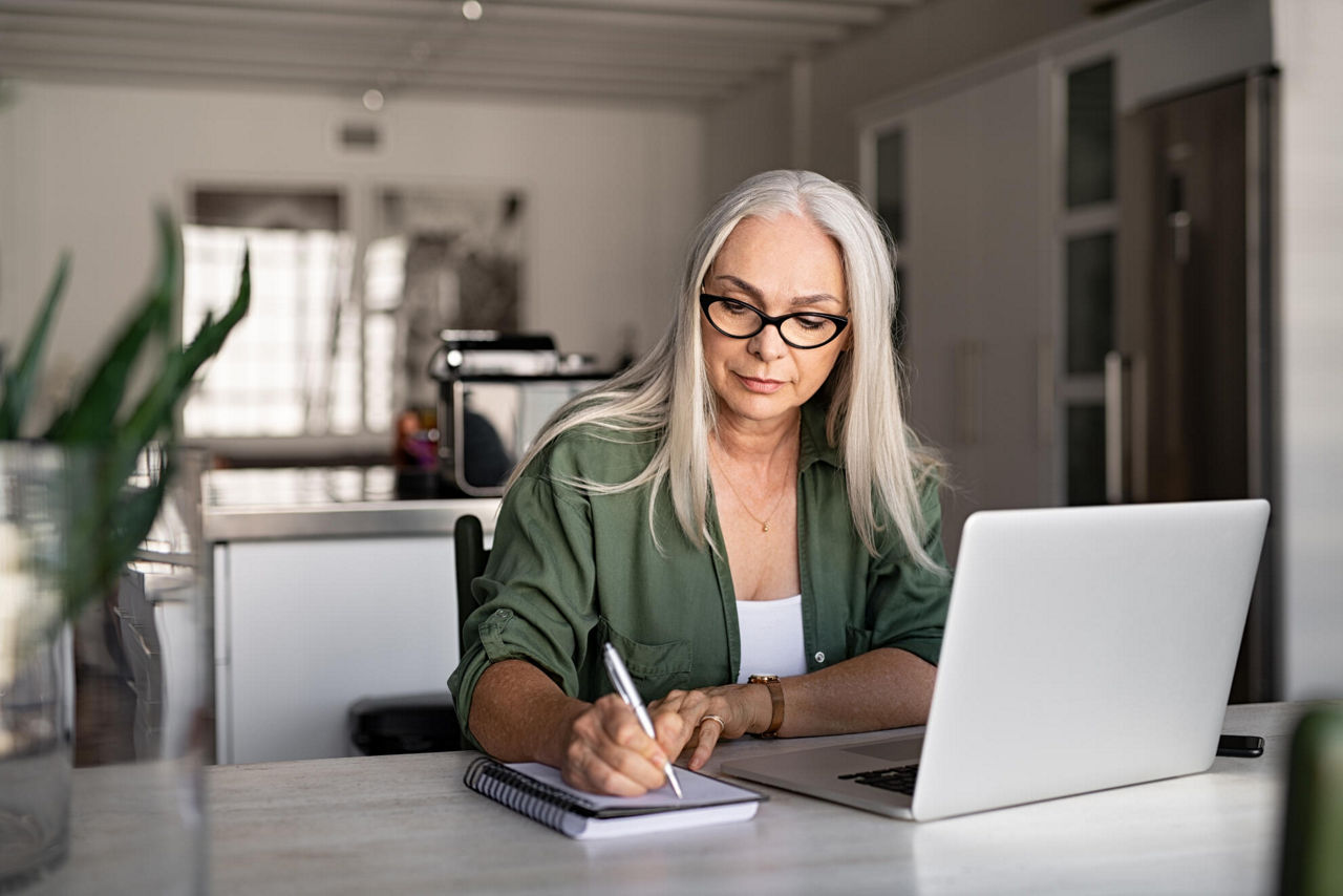 Eine ältere Dame sitzt zuhause vor ihrem Laptop und notiert etwas auf einen Zettel