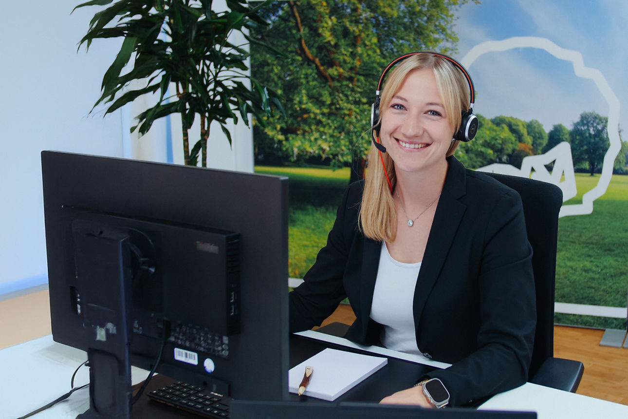 Eine Frau in einem schwarzen Blazer und mit Headset sitzt in einem Callcenter