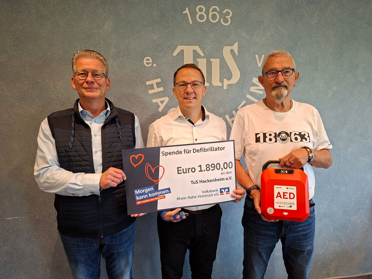 Drei Erwachsene stehen vor einer Wand halten einen Defibrillator und einen Spendenscheck in der Hand.