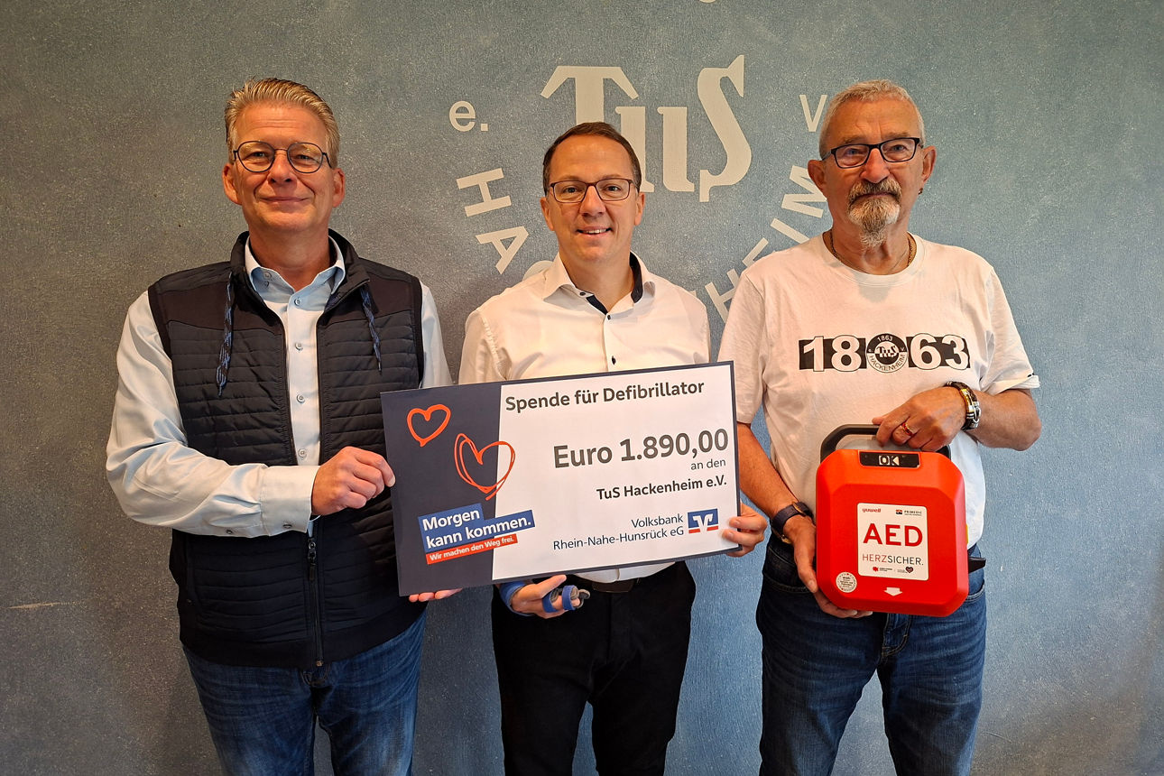 Drei Erwachsene stehen vor einer Wand halten einen Defibrillator und einen Spendenscheck in der Hand.