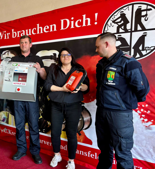 zwei Männer und eine Frau stehen vor einem Plakat und halten den Defibrillator in den Händen