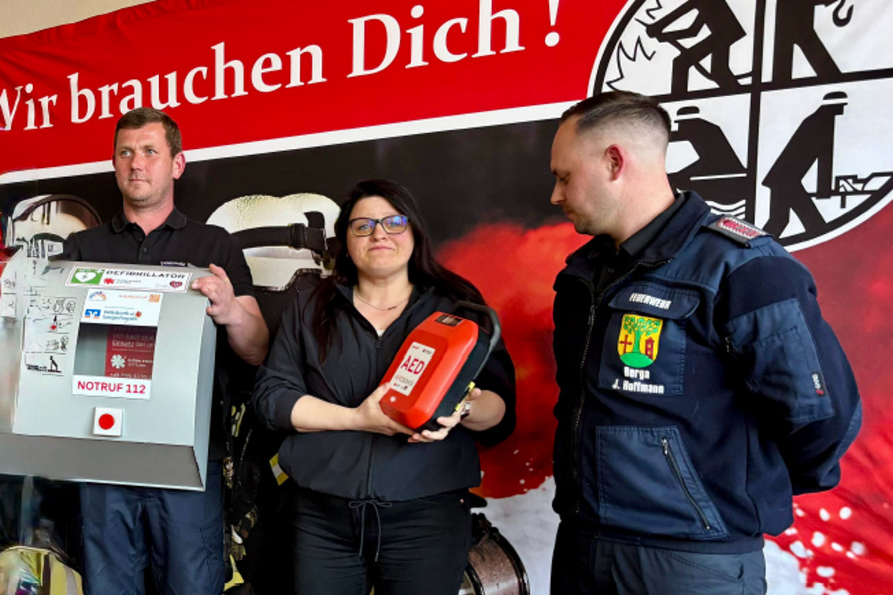 zwei Männer und eine Frau stehen vor einem Plakat und halten den Defibrillator in den Händen