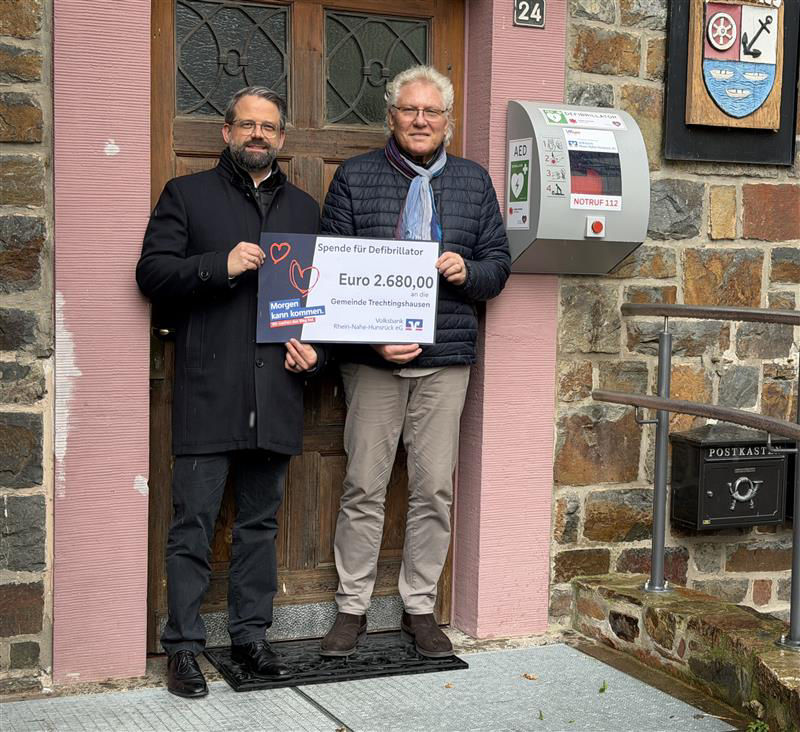 Zwei Erwachsene halten den Spendenscheck in der Hand und stehen vor einer Tür.