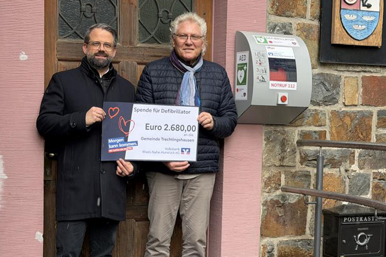 Zwei Erwachsene halten den Spendenscheck in der Hand und stehen vor einer Tür.