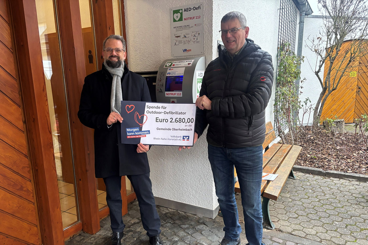 Zwei Erwachsene halten den Spendenscheck in der Hand und stehen vor einem Defibrillator.