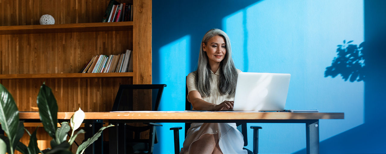 Frau mit grauen langen Haaren sam Laptop vor blauer Wand