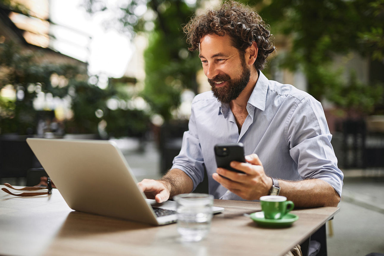 Mann sitzt im Cafe an Laptop und hat sein Handy in der Hand.
