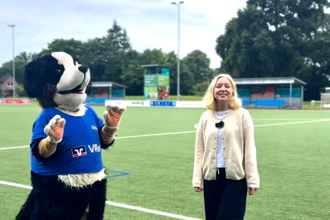 Eine junge Frau steht zusammen mit dem VReG-Maskottchen Anton auf einem Fußballfeld.