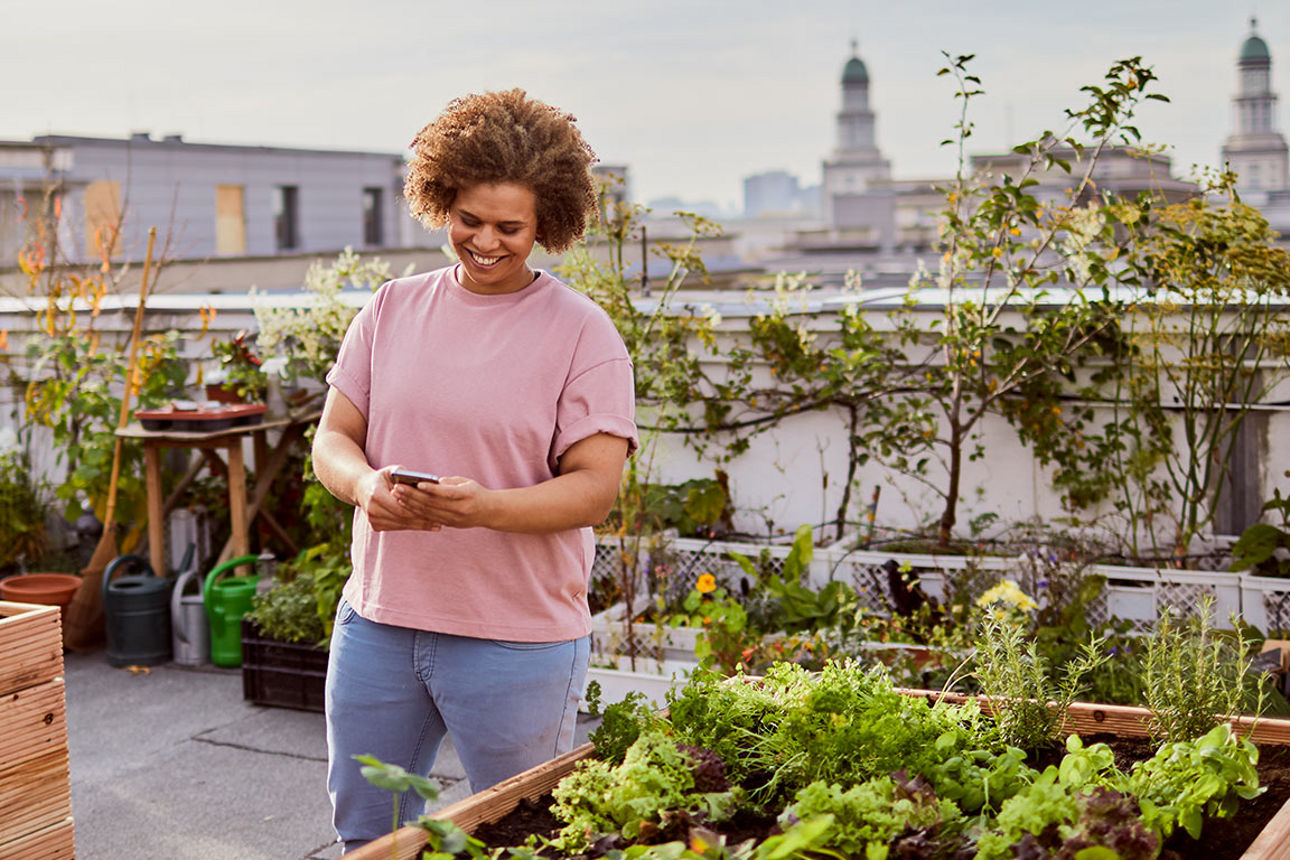Eine Frau steht auf einer Dachterrasse zwischen Hochbeeten und tippt lächelnd etwas in ihr Handy.