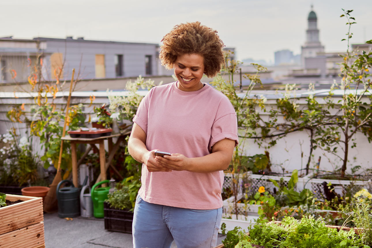 Eine Frau steht auf einer Dachterrasse zwischen Hochbeeten und tippt lächelnd etwas in ihr Handy.