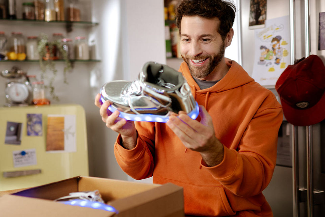 Ein junger Mann freut sich beim Paket auspacken über einen Sneaker mit leuchtender Sohle.