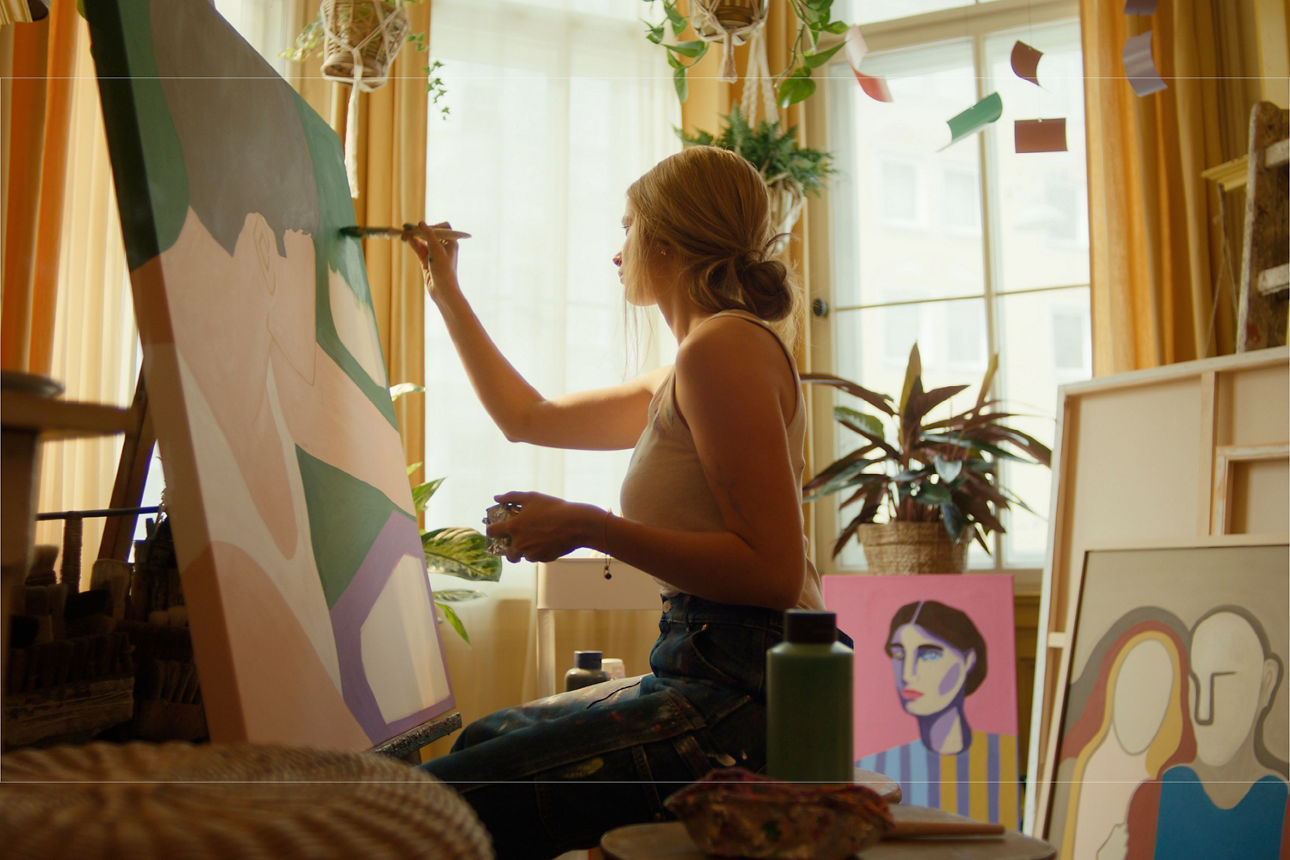 Eine junge Künstlerin sitzt vor einer Staffelei und malt in ihrem Atelier ein Bild. Darauf ist eine Person mit kurzen Haaren und weißen Top seitlich von oben zu sehen.
