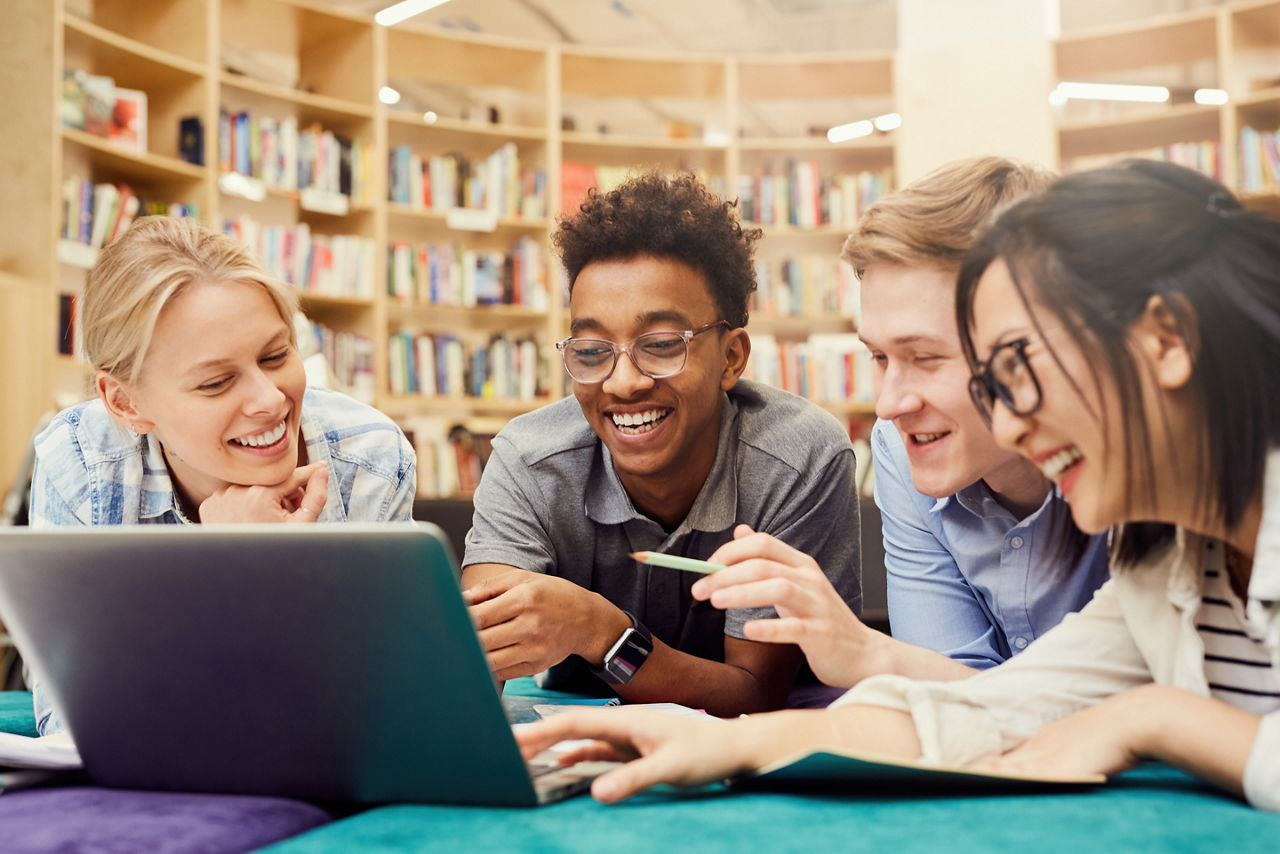 Studenten sitzen zusammen vor einem Laptop und lachen gemeinsam in der Bibliothek