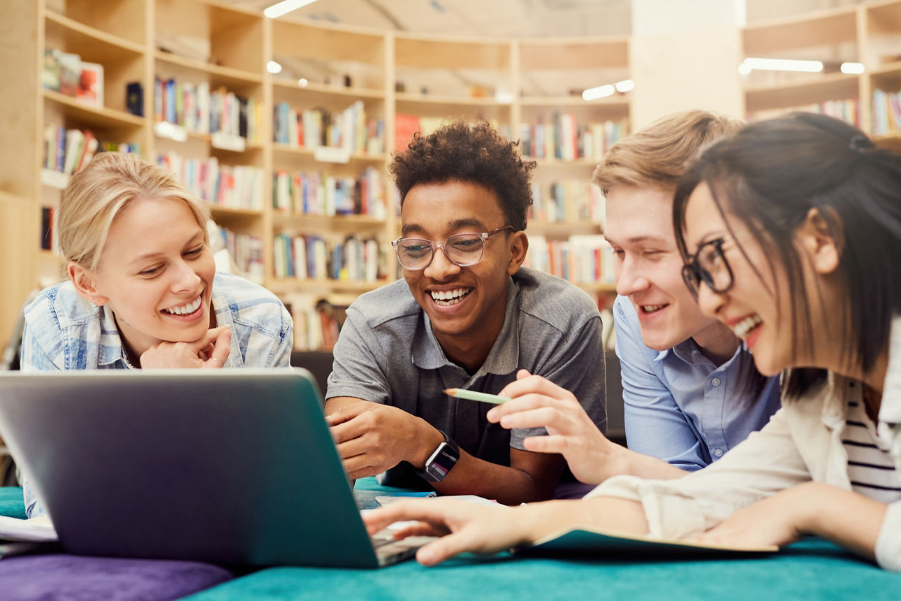 Studenten sitzen zusammen vor einem Laptop und lachen gemeinsam in der Bibliothek