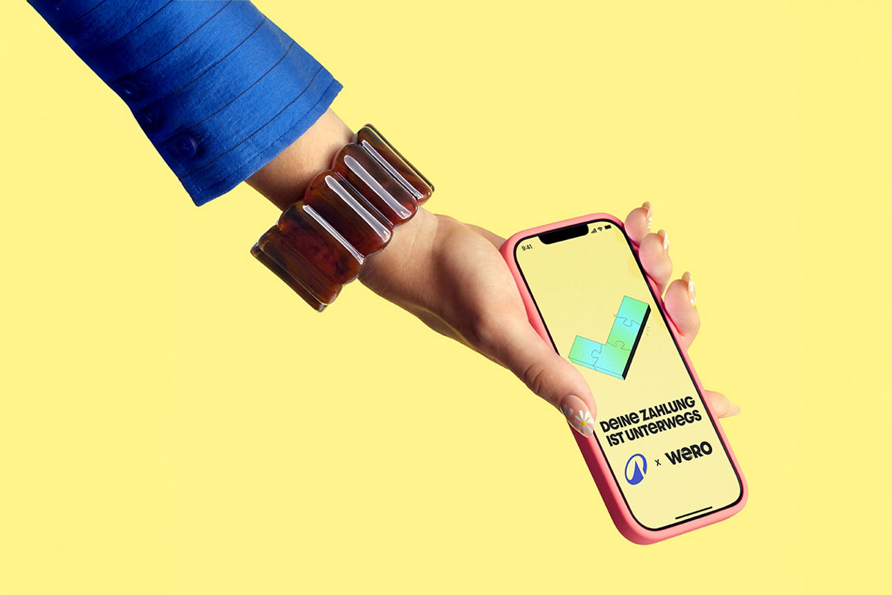 Frauenhand mit großem braunem Armreif hält ein Smartphone mit dem Text "Deine Zahlung ist unterwegs" mit den Logos von Wero und Decathlon, vor einem hellgelben Hintergrund.