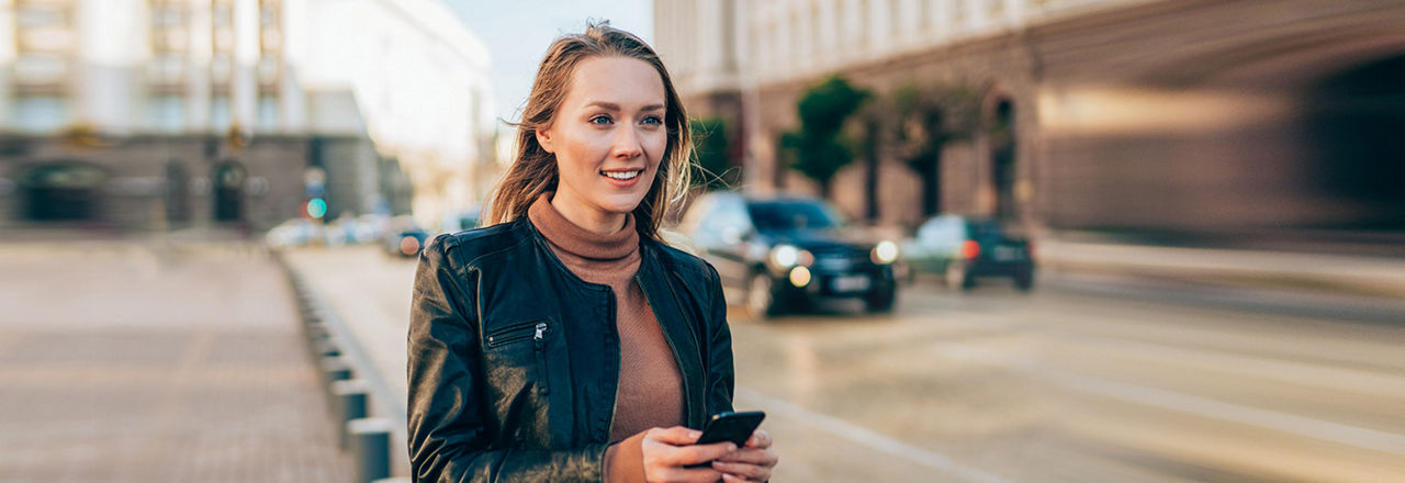 Eine junge Frau mit langen blonden Haaren blickt lächelnd nach rechts und hält ein schwarzes Smartphone in den Händen