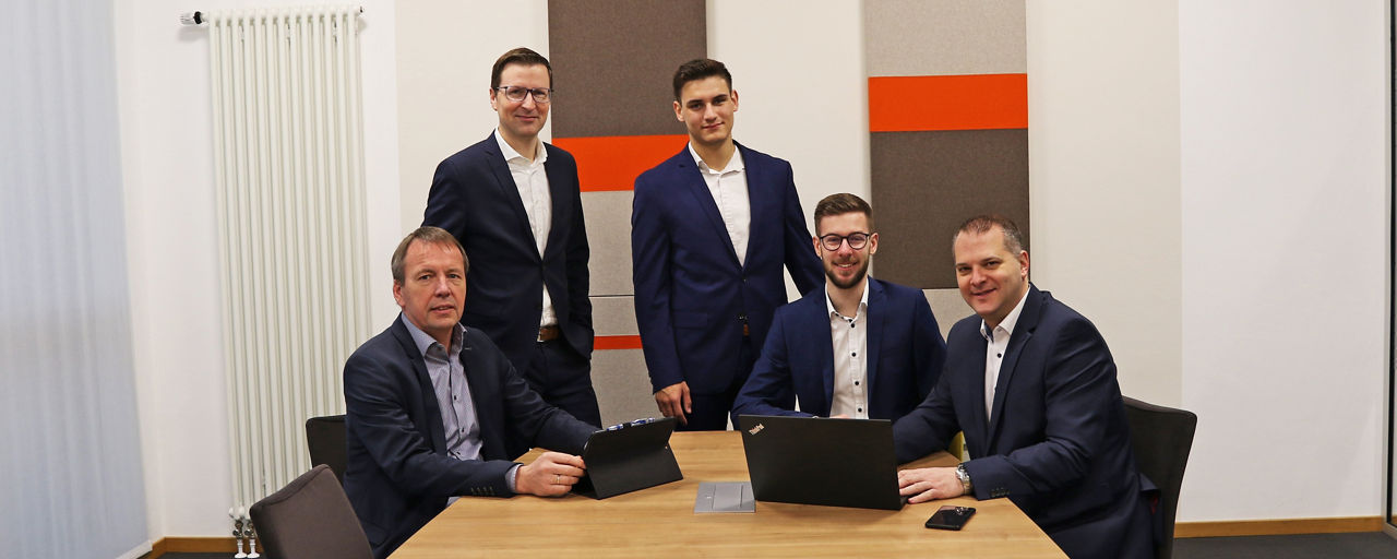 eBusiness Teamfoto mit fünf Männern im Konferenzraum vor farbigen Wandpaneelen.