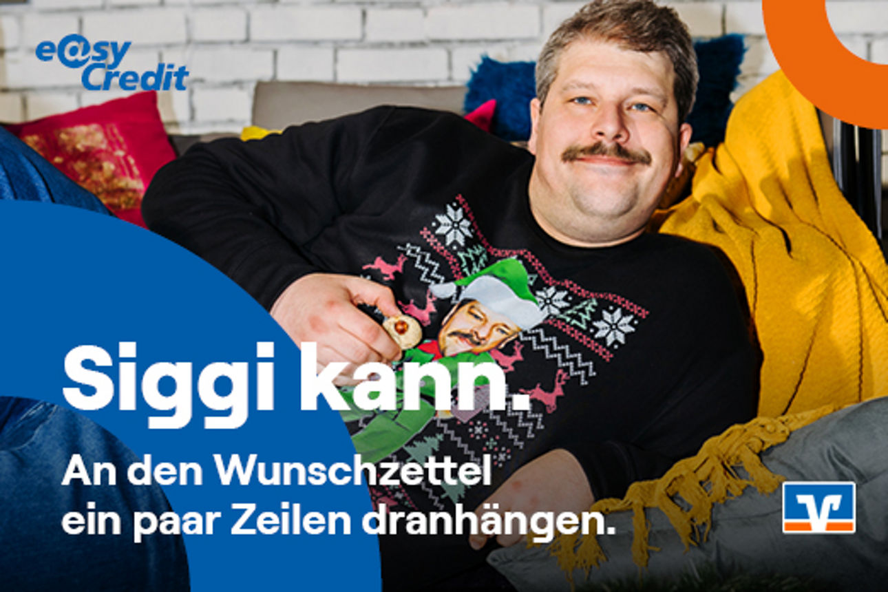 Mann im schwarzen Weihnachtspullover liegt zischen Geschenken und lächelt in die Kamera