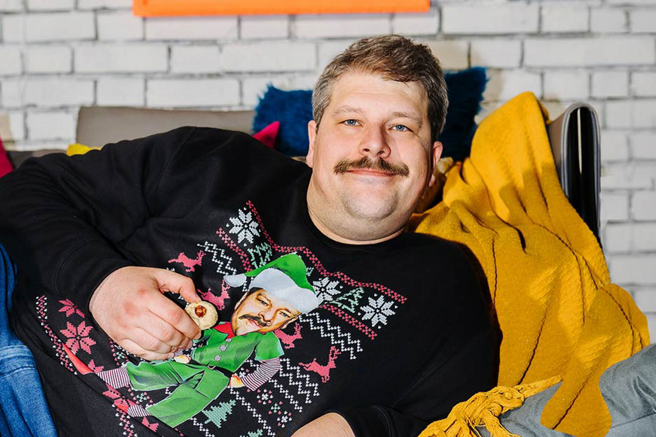 Mann mit Helm und orangefarbener Jacke sitzt auf einem blauen Moped und lächelt in die Kamera 