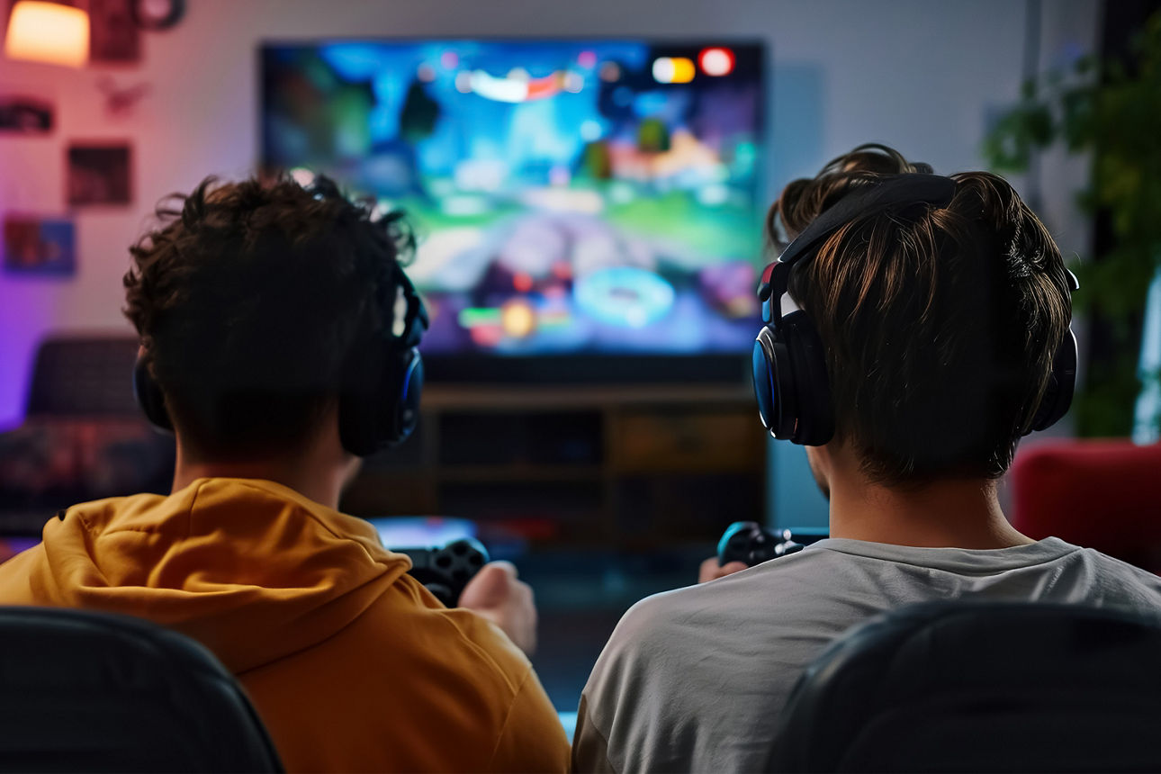 Zwei Personen sitzen nebeneinander, tragen Kopfhörer und spielen mit Controllern ein Videospiel auf einem großen Bildschirm.