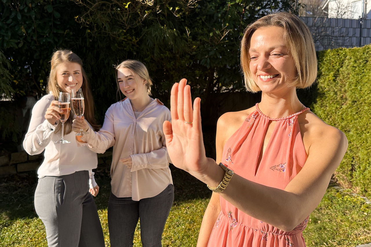 Junge Frau, die ihren Verlobungsring betrachtet; im Hintergrund: zwei Frauen, die mit Sekt anstoßen