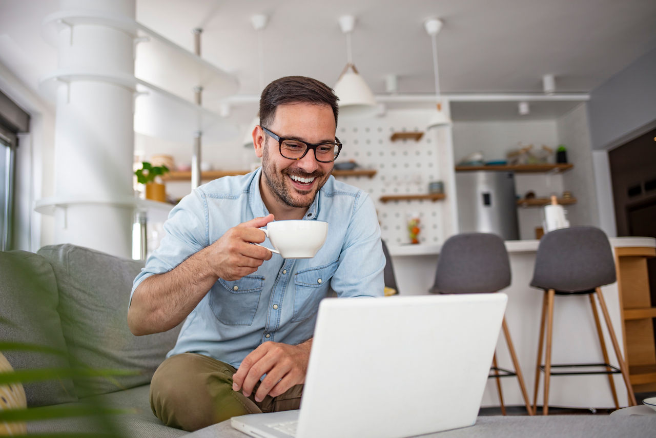 Mann sitzt mit einer Tasse in der Hand auf dem Sofa vor dem Laptop