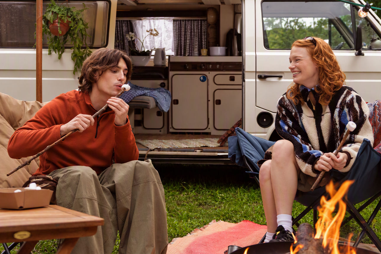 Frau und Mann sitzen am Lagerfeuer vor einem Campingwagen und grillen Marshmallows