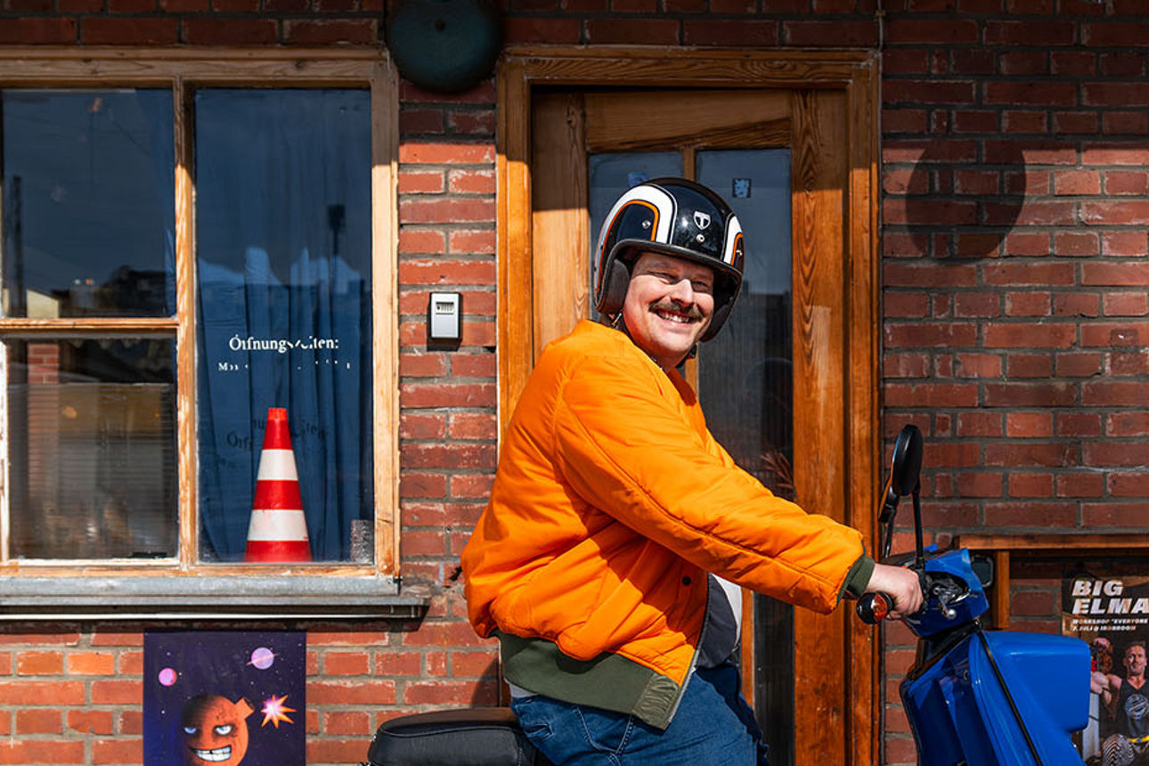 Lachender Mann in orangener Jacke auf blauem Roller