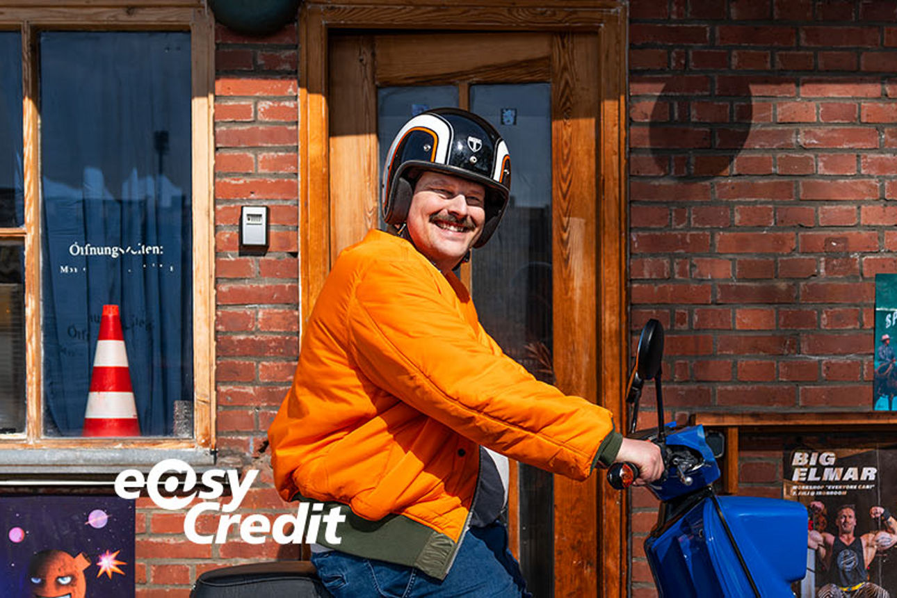 Ein Mann sitzt auf dem Moped vor einem Geschäft und lächelt in die Kamera.
