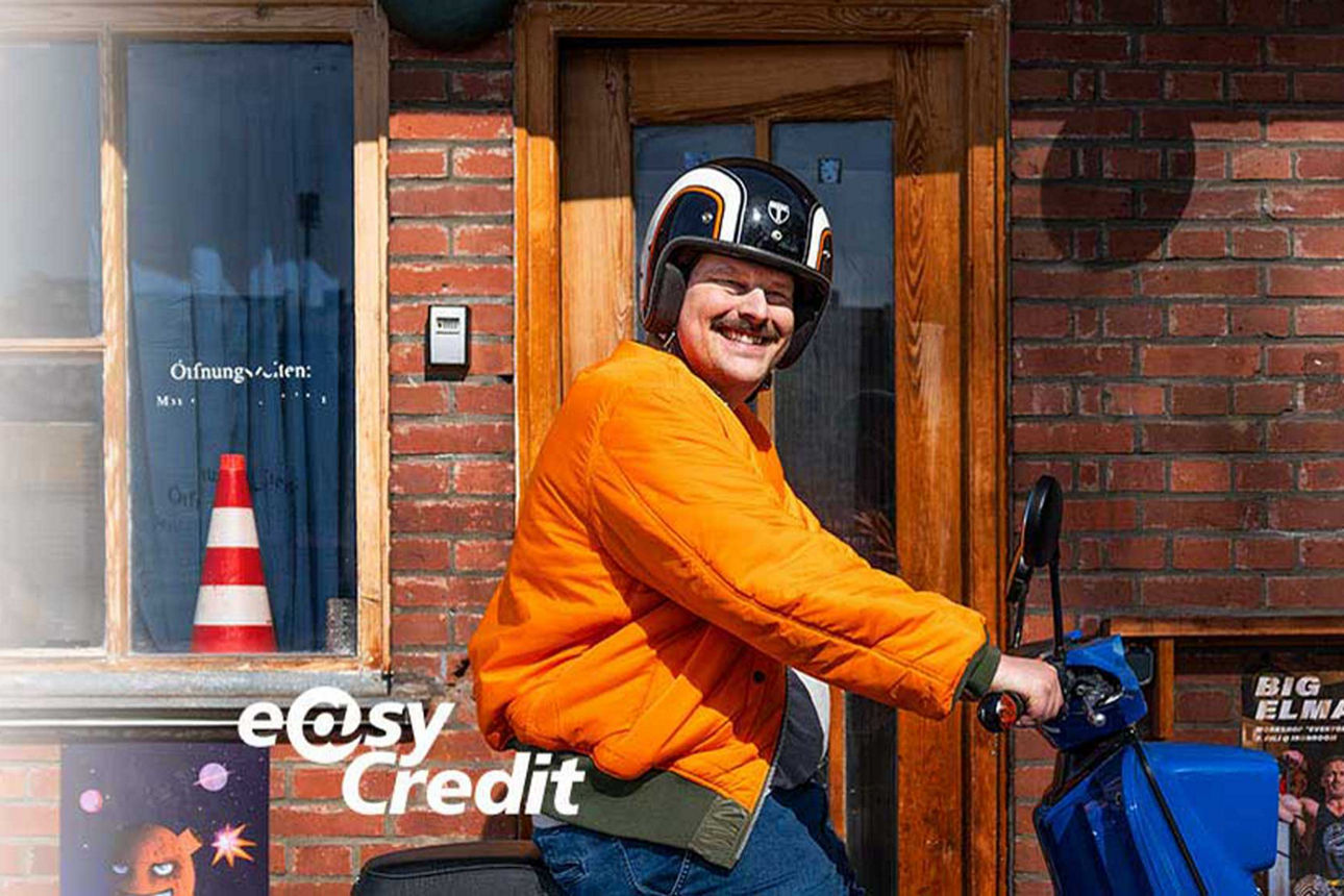 Ein Mann in orangenen Sweatshirt liegt auf dem Rasen, spielt auf einem kleinen Klavier und schaut in den Himmel