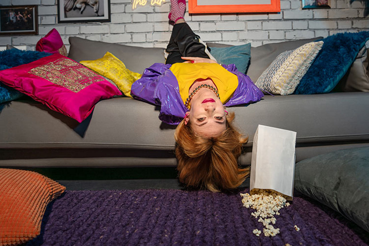 Eine Frau liegt kopfüber auf dem Sofa zwischen bunten Kissen und blickt nach vorn.