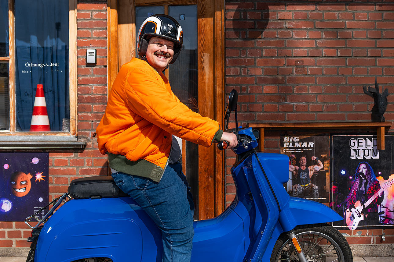 Mann mit Helm und orangefarbener Jacke sitzt auf einem blauen Moped und lächelt in die Kamera 