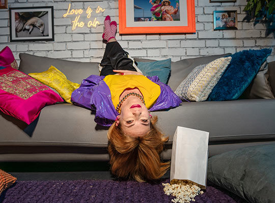 Eine Frau liegt mit dem Kopf nach unten auf der Couch, ihre Füße liegen über der Rückenlehne. Eine Tüte Popcorn fällt zu Boden