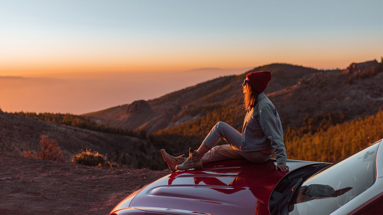 Junge Frau sitzt auf Motorhaube Ihres roten Autos und genießt den Sonnenaufgang
