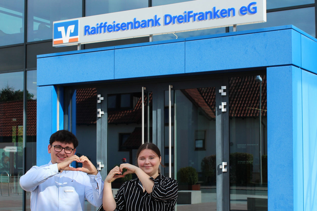 Zwei Azubis stehen vor dem Hauptgebäude in Heßdorf. Ihre Hände formen je ein Herz.