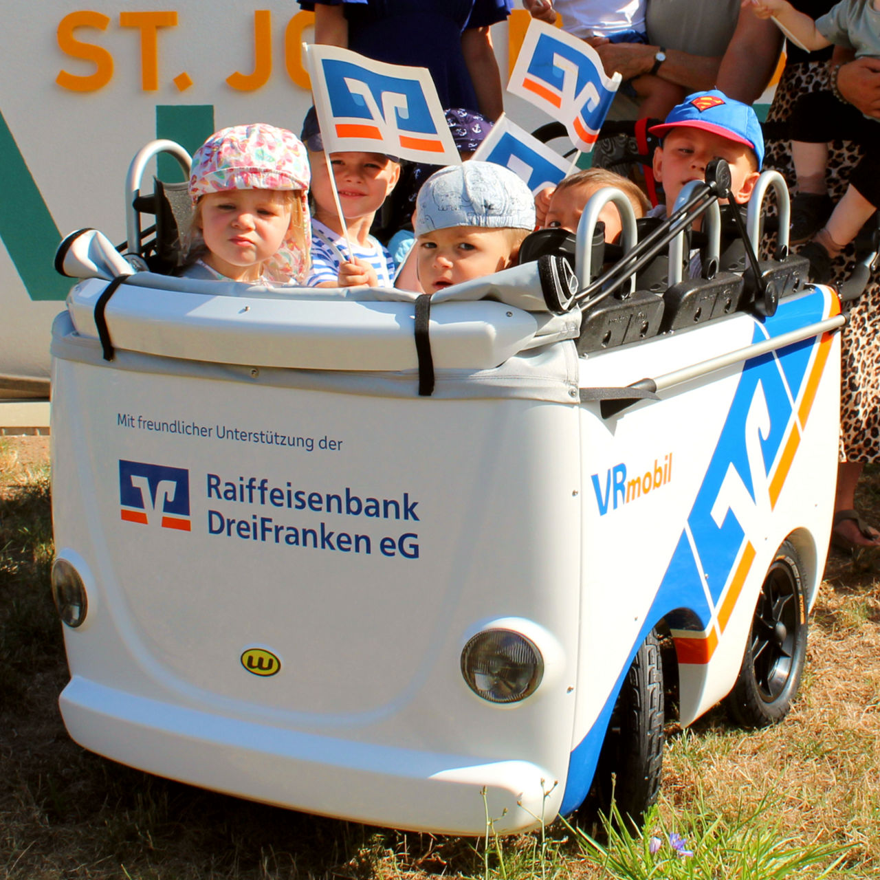 Kinder sitzen im Kinderbus und halten Fahnen mit dem VR-Logo in den Händen.