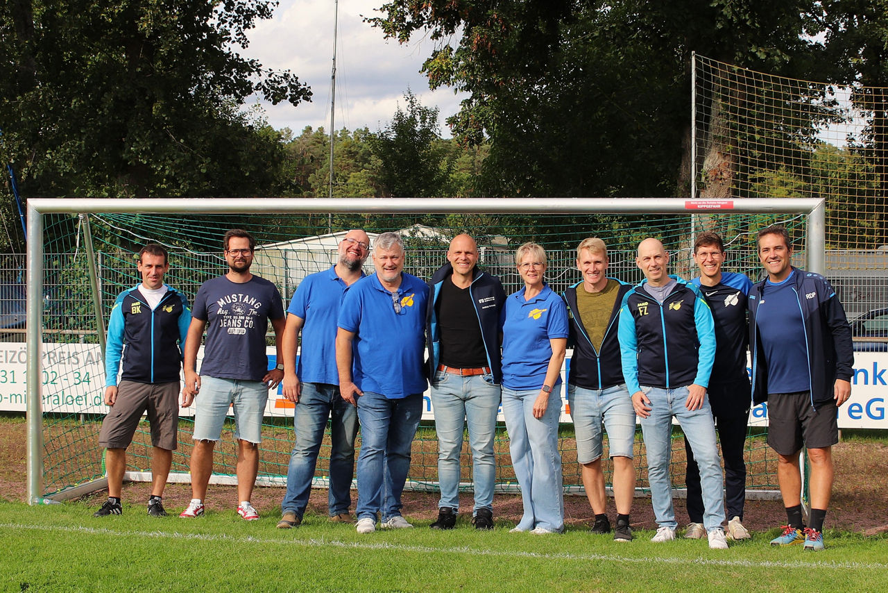 Vereinsmitglieder strahlen mit Bankvorstand Andreas Heilmann auf einem Fußballplatz in die Kamera