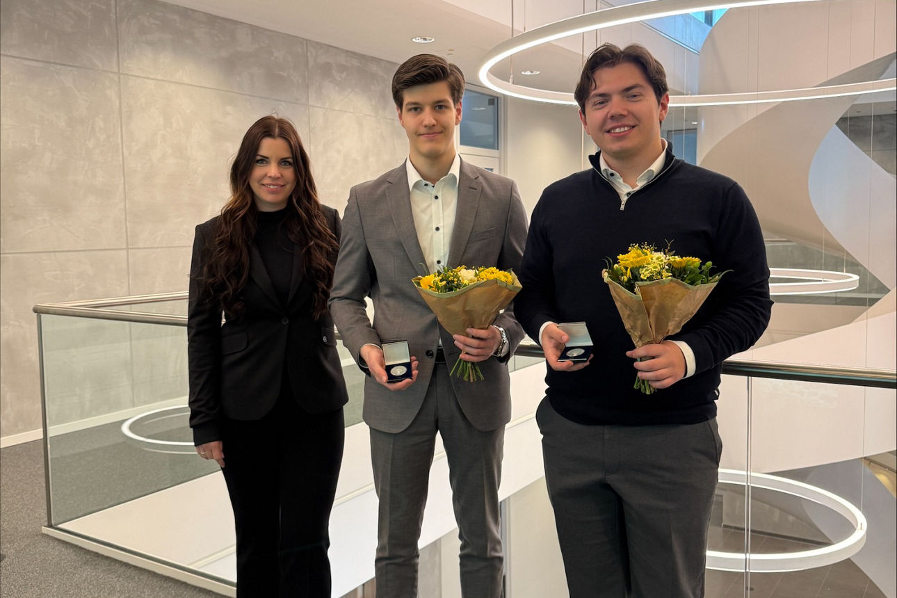Frau und zwei Männer stehen in einem großen Gebäude und halten Blumen sowie ein kleines Geschenk.