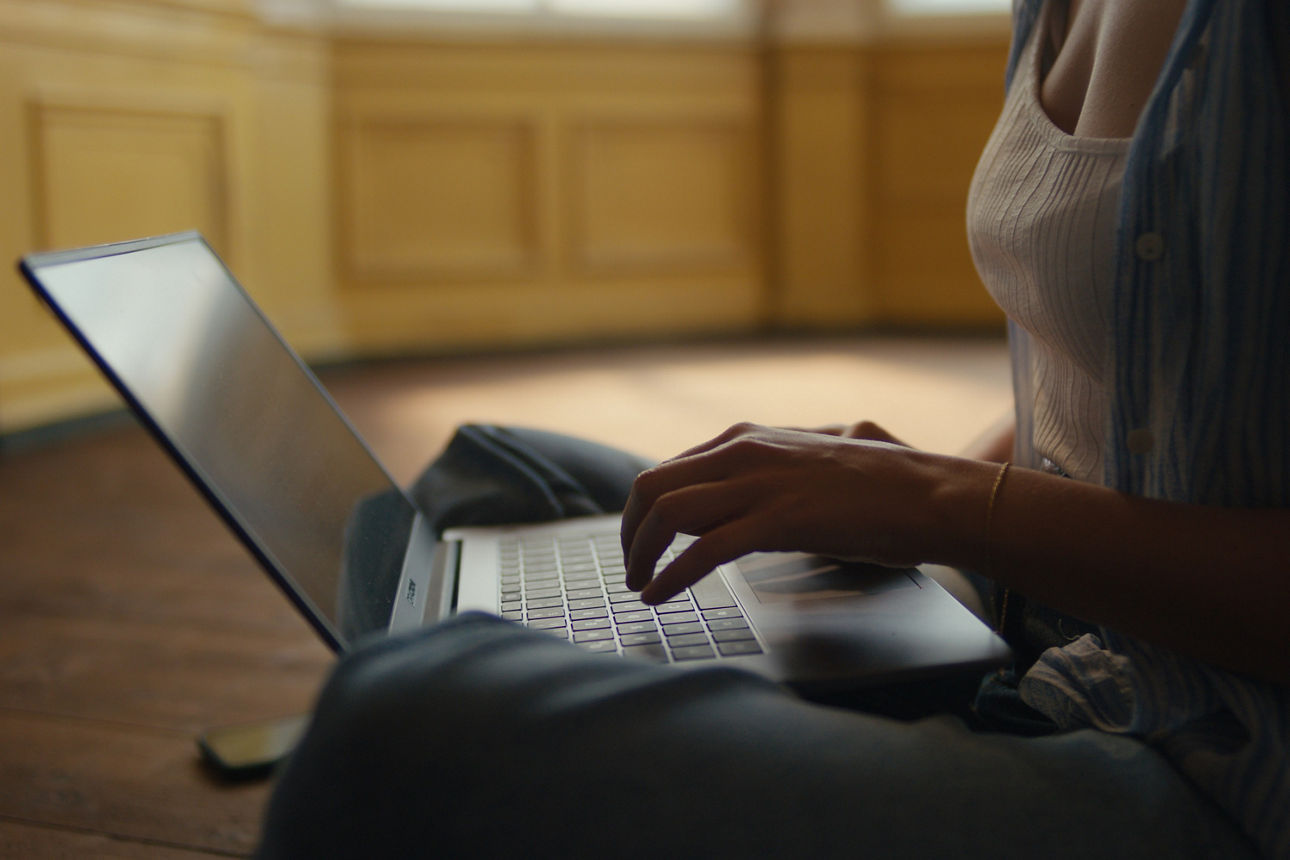Man sieht den Oberkörper einer Frau. Auf ihrem Schoß ist ein geöffneter Laptop, auf dem sie tippt.