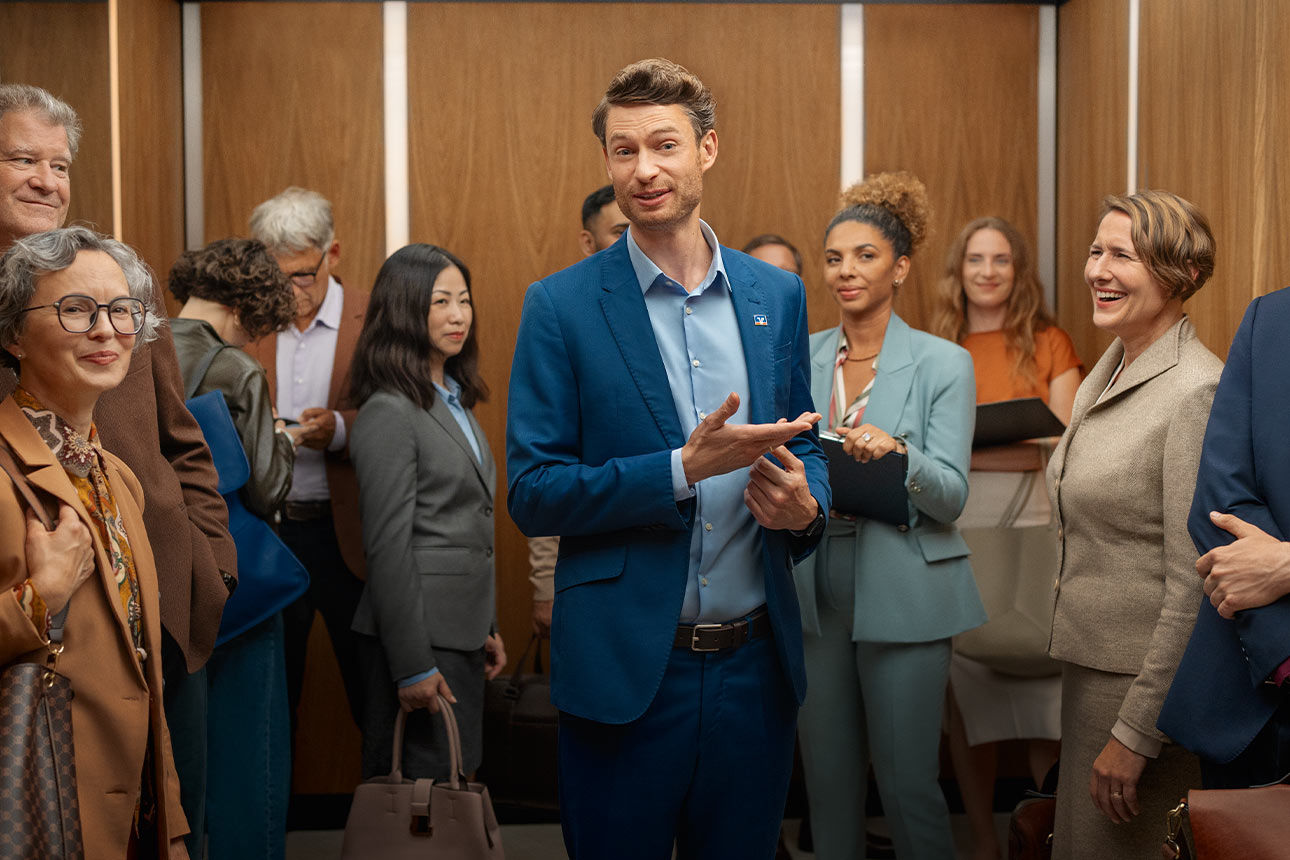 Ein Mann im Anzug steht umgeben von anderen Menschen in Geschäftskleidung in einem Aufzug