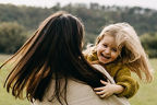Baby berührt mit ausgestrecktem Arm das Gesicht der Mutter mit lockigem Haar vor unscharfem Hintergrund