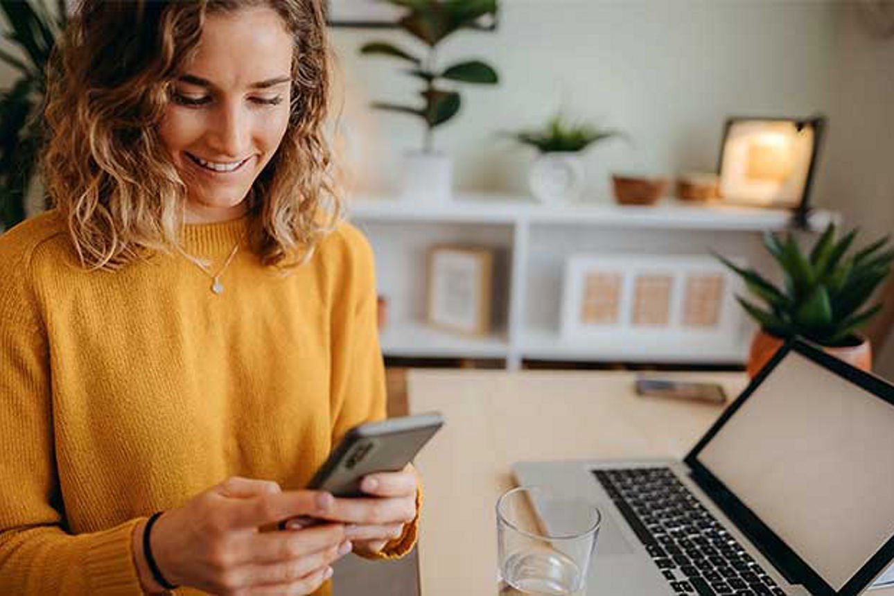 Junge Frau schaut lächeln auf ihr Smartphone, daneben ein geöffneter Laptop auf dem Schreibtisch