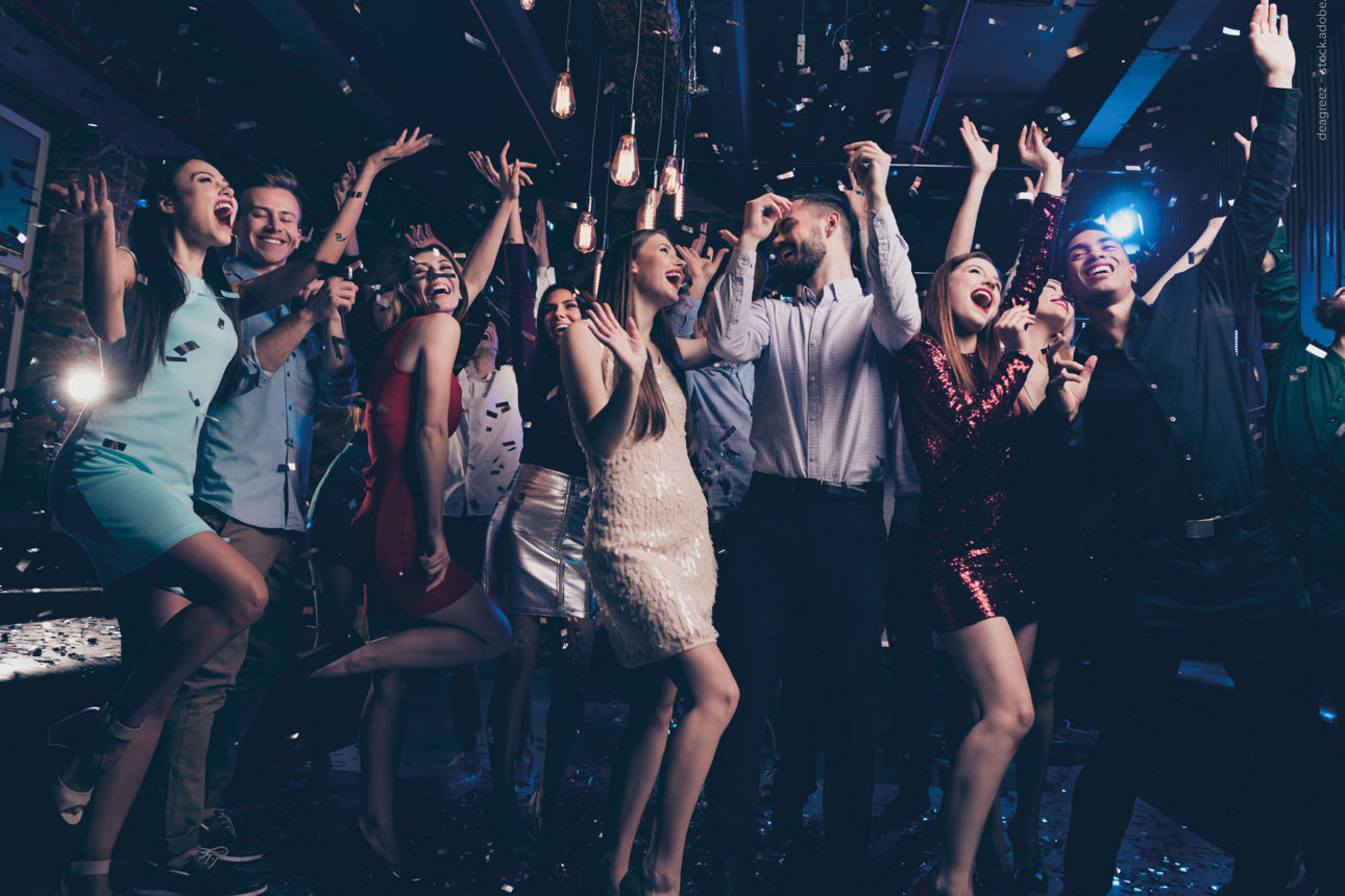Junge Menschen tanzen fröhlich jubelnd mit hoch gehobenen Händen in einem abgedunkelten Partyraum