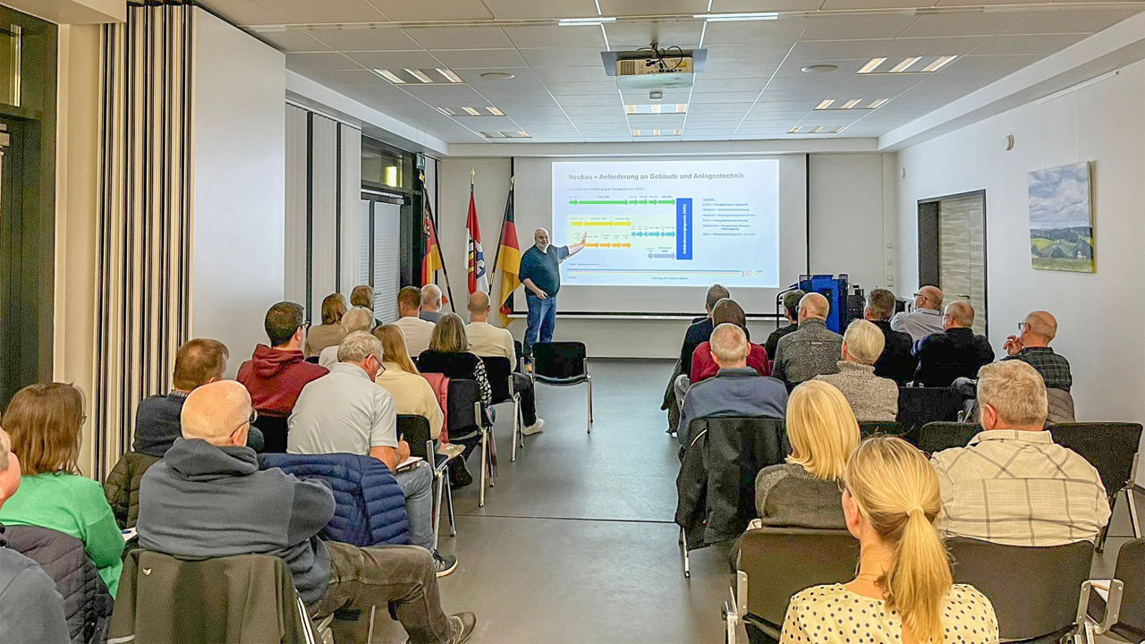 Eine Gruppe von Leuten sitzt in einem Saal vor einer Leinwand, auf der ein Mann eine Präsentation zeigt
