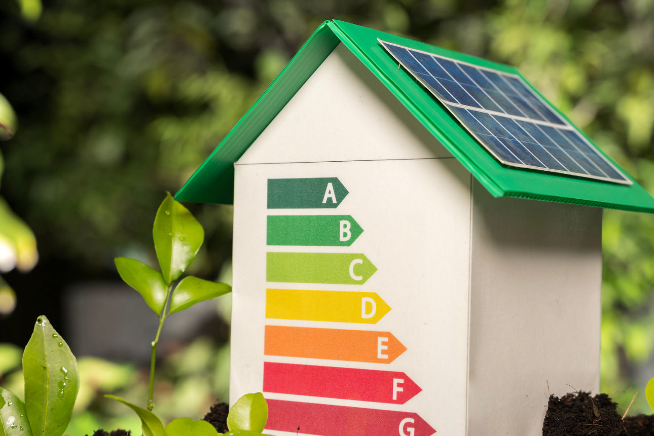 Modellhaus mit Solardach und farbiger Energieeffizienzskala steht auf Erde zwischen jungen Pflanzen.
