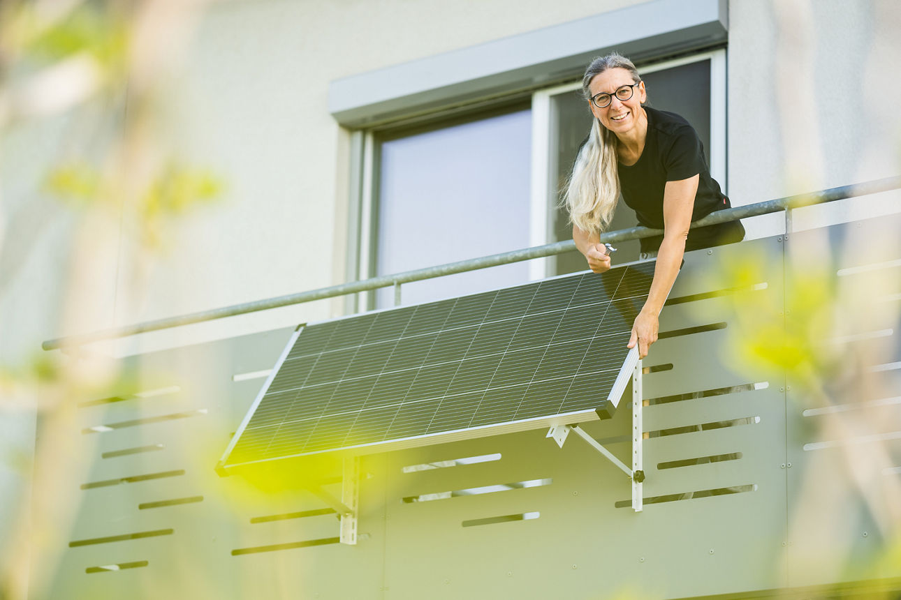 Person montiert ein Solarmodul am Balkon eines Wohnhauses, im Vordergrund unscharfe grüne Blätter.