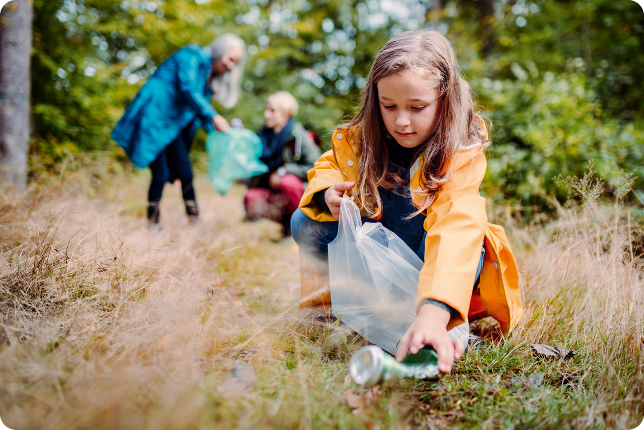 Ein Mädchen sammelt im Wald Müll in eine Plastiktüte, während zwei weitere Personen im Hintergrund ebenfalls aufräumen.