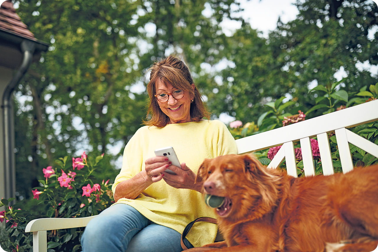 Eine Frau sitzt mit ihrem Hund auf einer Bank und schaut lächelnd auf ihr Smartphone