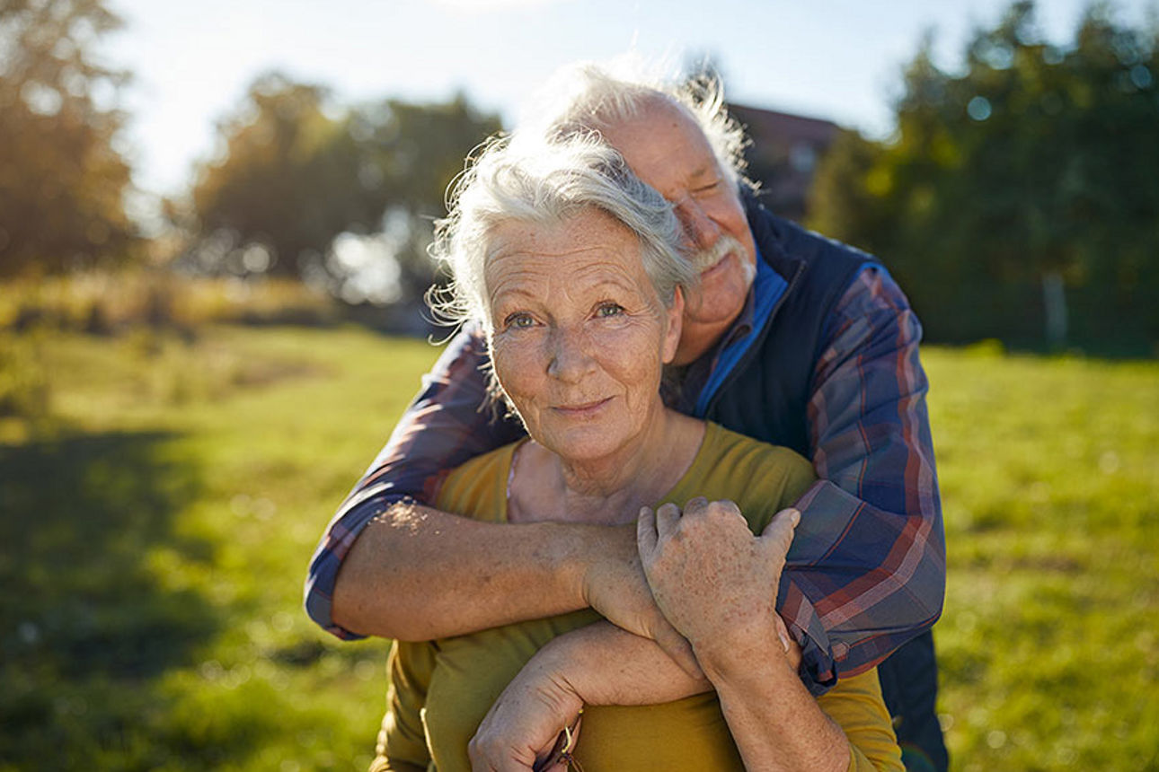 Älterer Herr mit grauen Haaren umarmt seine Frau von hinten